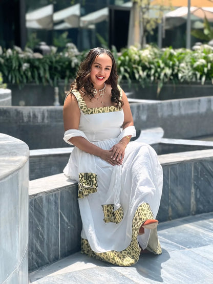 Woman in a white and gold traditional outfit sitting outdoors with plants in the background