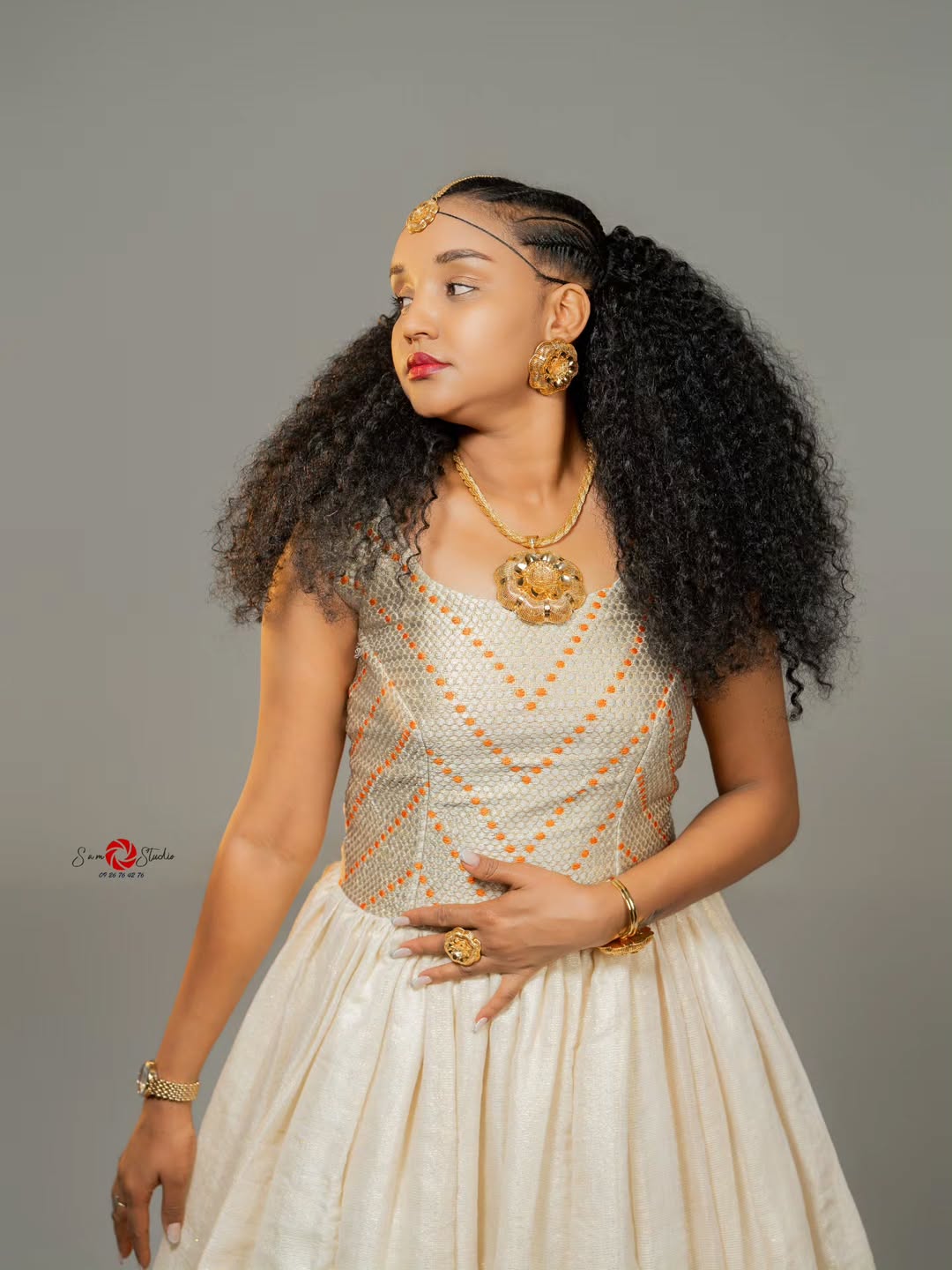 Woman in traditional habesha attire with gold jewelry against a gray background