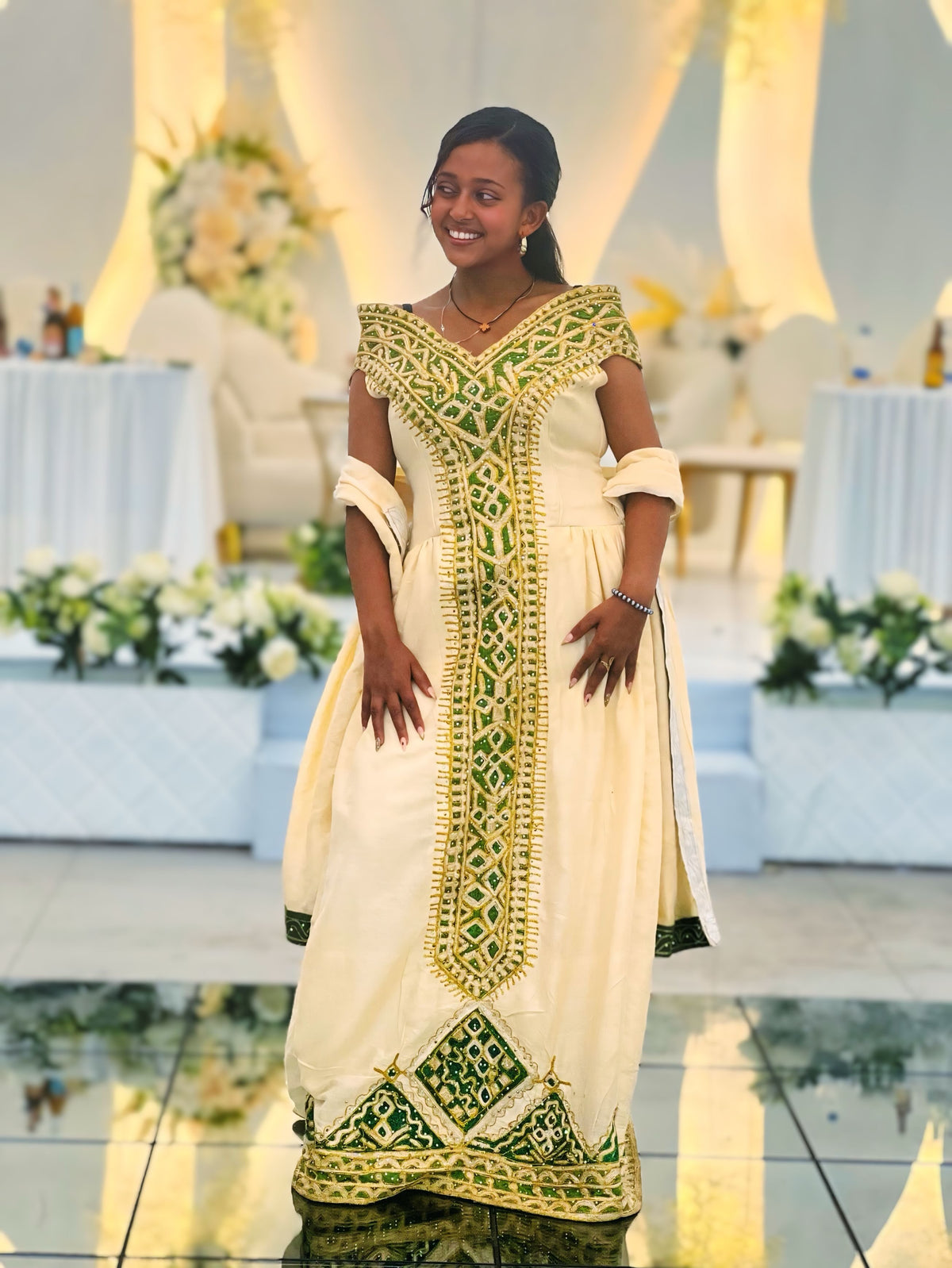 A woman wearing a cream-colored traditional Ethiopian National Dresses with intricate green and gold embroidery, standing in a decorated event hall with tables and floral arrangements in the background.