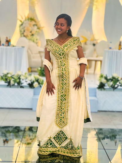 A woman wearing a cream-colored traditional Ethiopian National Dresses with intricate green and gold embroidery, standing in a decorated event hall with tables and floral arrangements in the background.