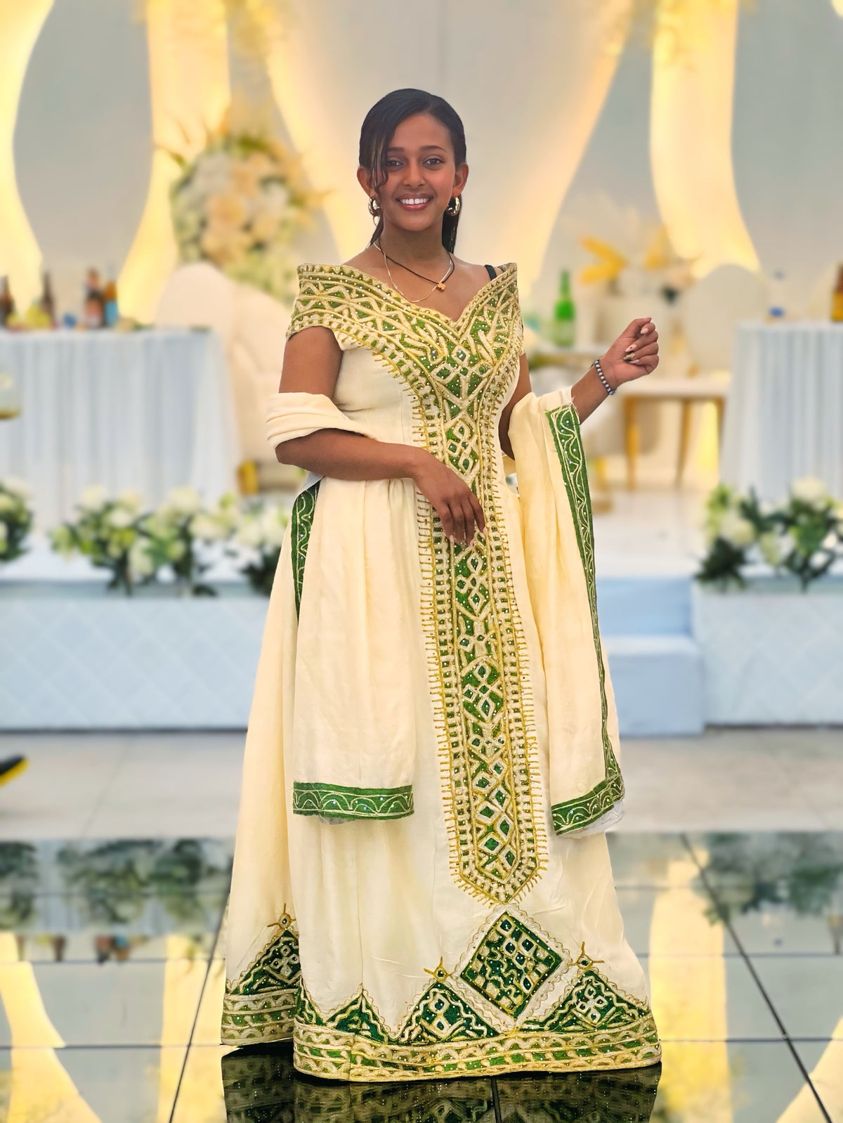 A woman stands in a decorated event hall wearing a cream-colored traditional Ethiopian National Dresses with extensive green and gold embroidery. The dress features an off-the-shoulder neckline, a draped dupatta, and detailed geometric patterns along the borders.