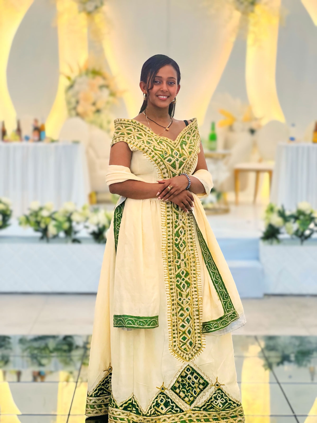 A woman wearing a traditional Indian salwar Ethiopian National Dresses in off-white with green and gold embroidery, standing in a decorated event hall.