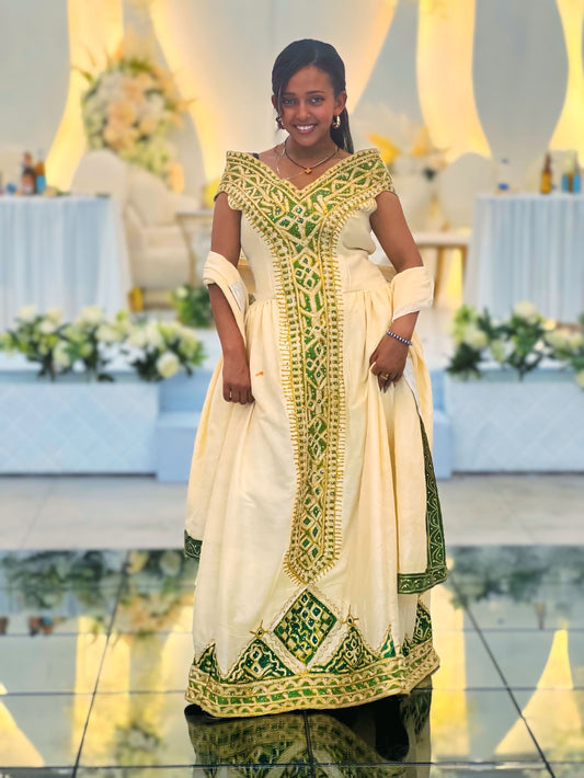 A woman stands on a glossy floor wearing a traditional long Ethiopian National Dresses with an ivory base and green and gold embroidered patterns. The dress features a V-neck, short sleeves, and a full skirt with ornate border detailing.