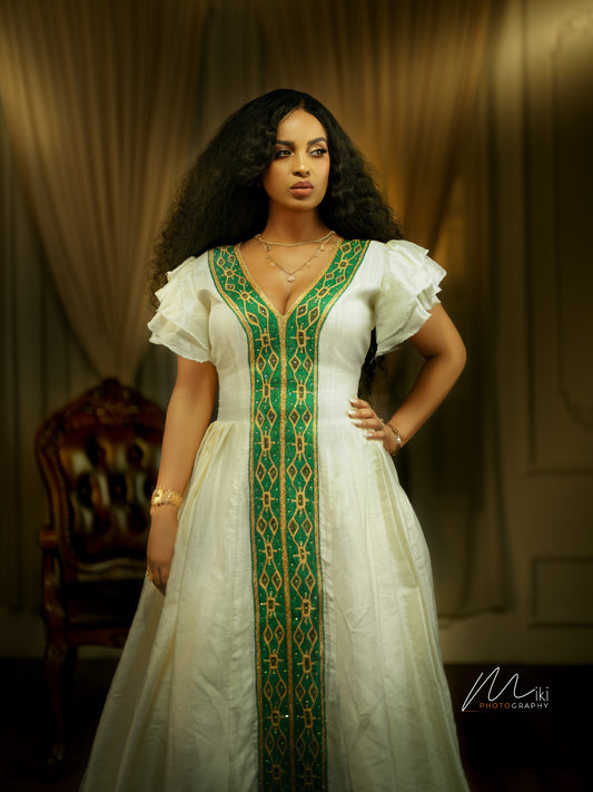 A woman modeling a white traditional Ethiopian dress with a prominent green and gold embroidered vertical panel down the front, ruffled short sleeves, and a deep V-neckline, set against a warm, dimly lit background with a dark wooden chair.