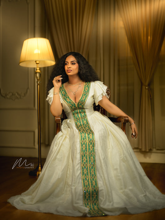 A woman seated in a chair wearing a voluminous white Ethiopian dress with a deep V-neck and a prominent green and gold patterned central panel. The setting is an elegant room with warm lighting, a floor lamp, and draped curtains.