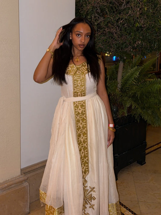 Woman wearing a white Habesha Kemis with gold embroidery indoors.