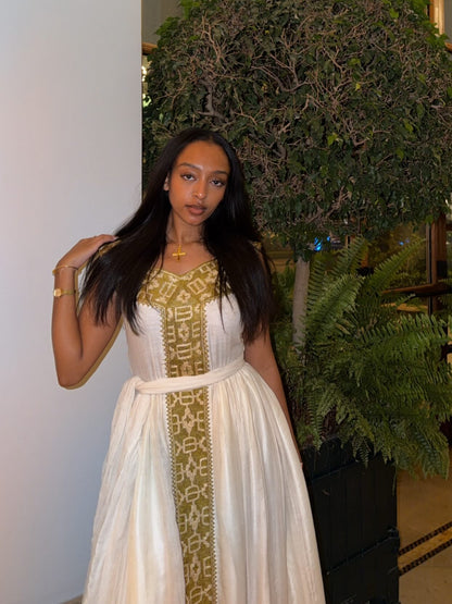 Woman in a white Habesha Kemis with gold embroidery standing next to a plant indoors.