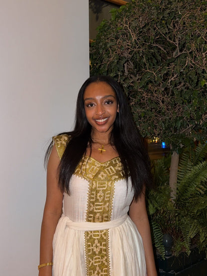 Woman in a white Habesha Kemis with gold embroidery standing in front of a green plant.