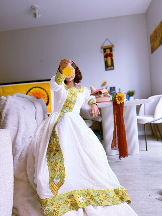 A woman wearing a long white Habesha Kemis  with ornate green and gold embroidery sits on a white sofa in a bright living room, holding a glass of orange juice.
