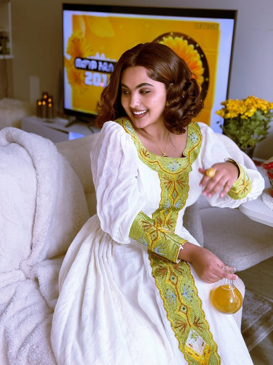A woman wearing a white Habesha Kemis  with green and gold embroidered details sits on a white couch, holding a small glass bottle. A television in the background displays a yellow graphic.