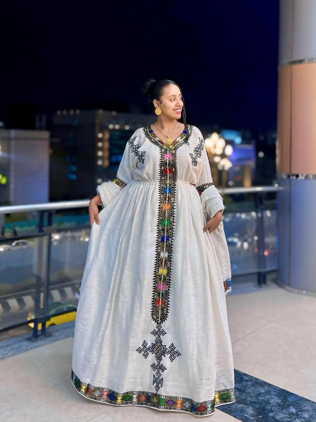 Woman in a long, embroidered Ethiopian Dress  standing on a rooftop at night.