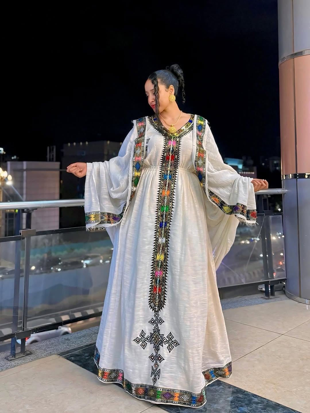 Woman in a traditional embroidered Ethiopian Dress  standing on a balcony at night.