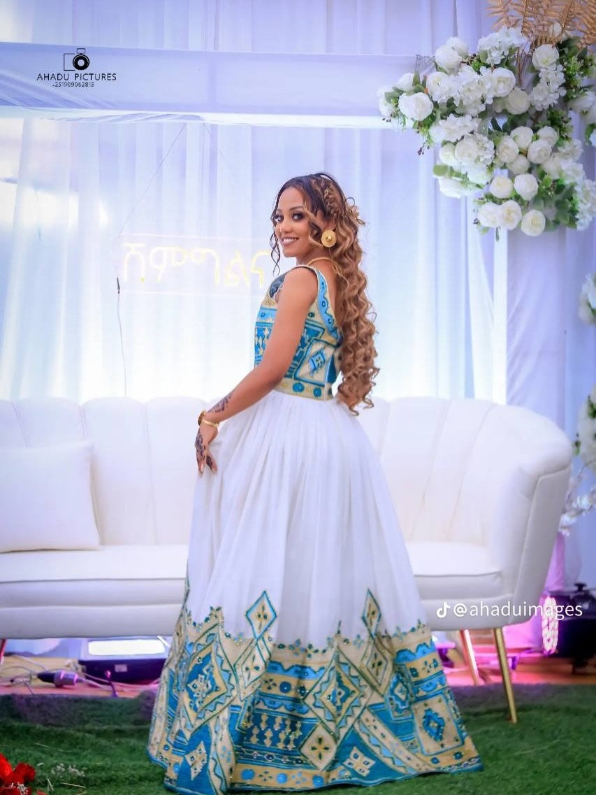 Woman in a traditional habesha wedding dress with white and blue patterns in a decorated room.
