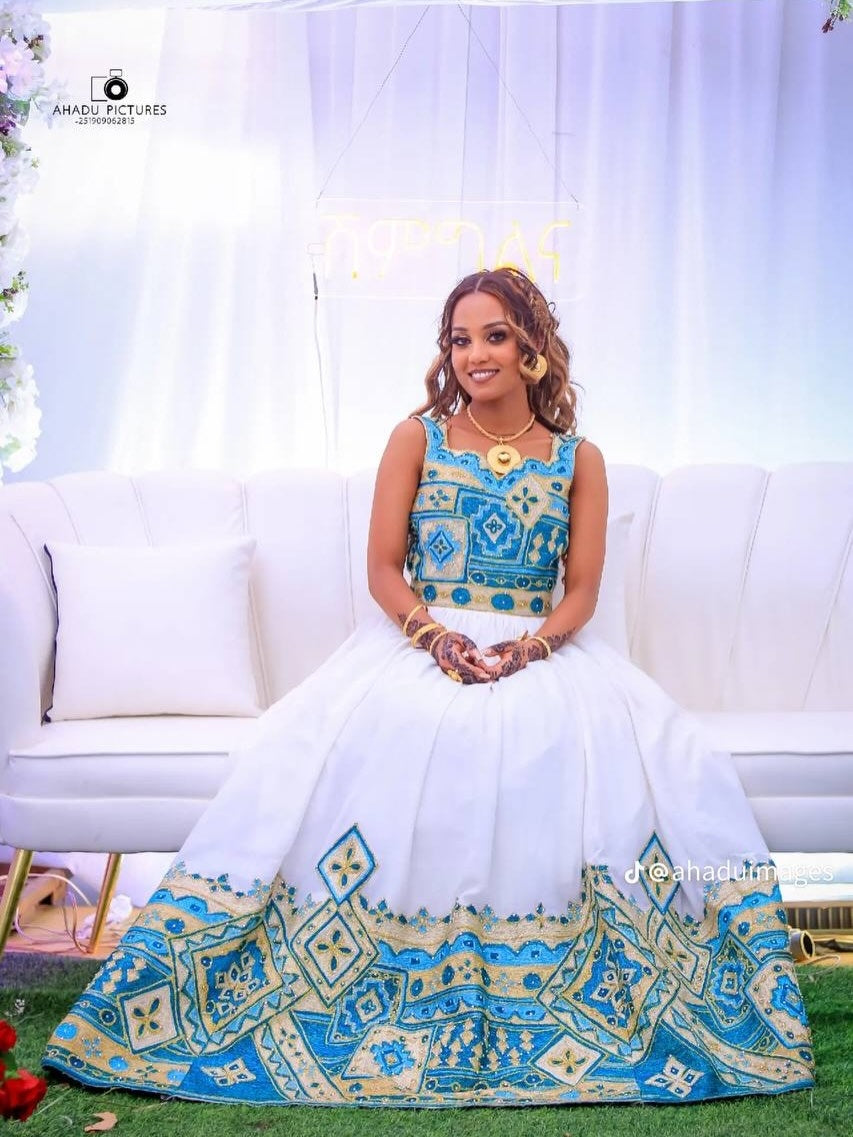 Woman in a blue and white traditional habesha wedding dress sitting on a white couch with floral decorations.