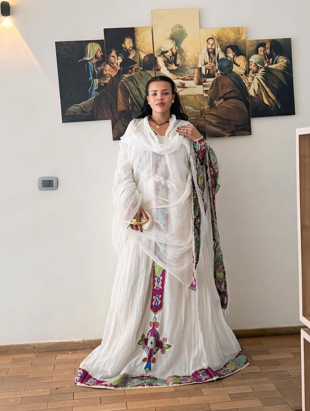 Woman in a white Purple Habesha Zuriastanding in front of religious paintings on a wall.