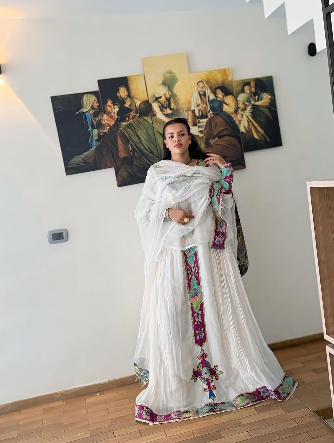 Woman in a Purple Habesha Zuria with colorful patterns standing in a room with classical paintings on the wall.