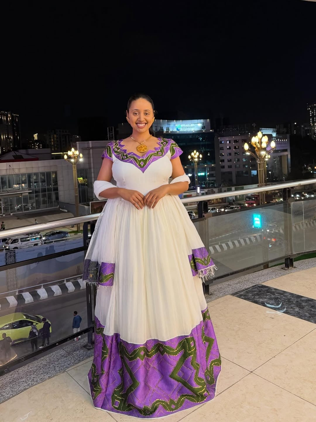Woman in a white and purple traditional outfit standing on a rooftop at night with city lights in the background.