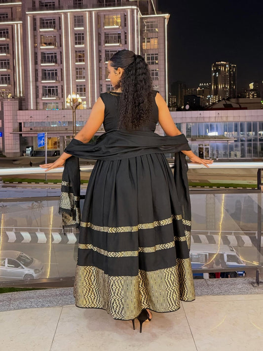 A woman stands on a balcony at night wearing a black traditional Habesha zuria with gold patterned borders and a draped black shawl. The background shows illuminated city buildings and a glass railing.