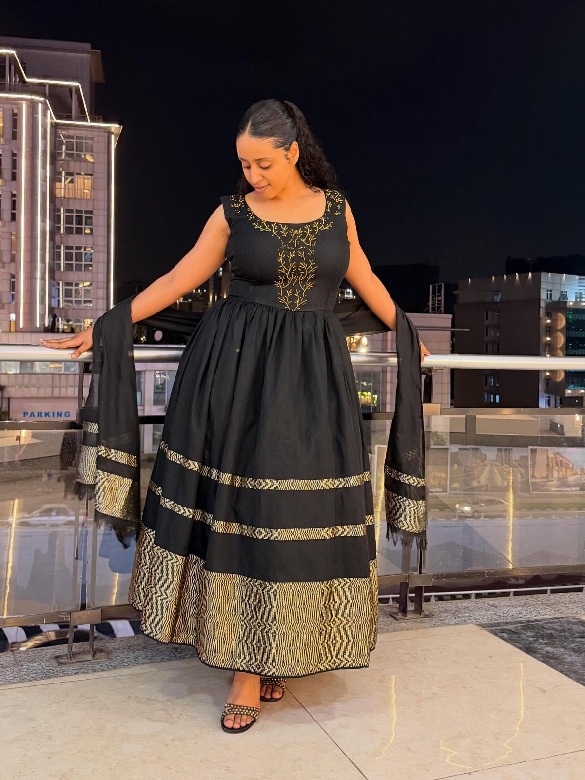 A woman modeling a black sleeveless maxi Habesha zuria with gold embroidery on the bodice and a wide gold patterned hem, paired with a matching dupatta, set against a nighttime city balcony background.