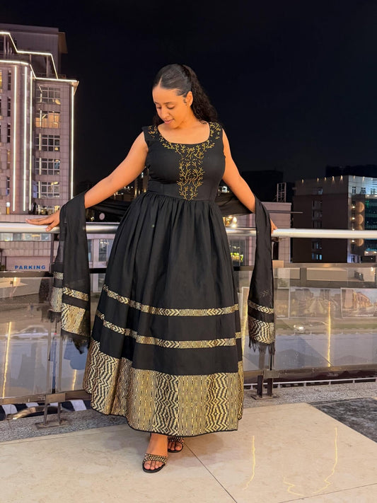 A woman modeling a black sleeveless maxi Habesha zuria with gold embroidery on the bodice and a wide gold patterned hem, paired with a matching dupatta, set against a nighttime city balcony background.