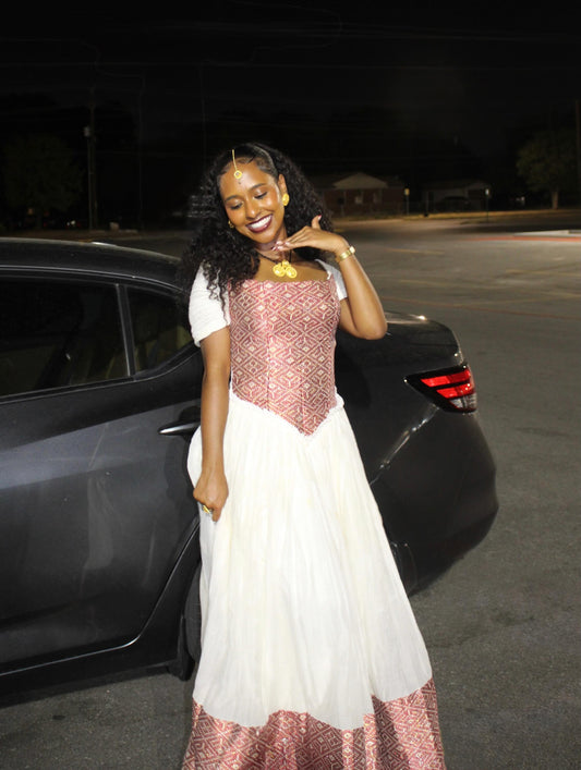 A woman posing in a white and pink Ethiopian Dress next to a black car at night.
