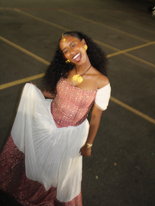 A woman posing outdoors at night in a white and metallic pink Ethiopian Dress, smiling broadly. She is wearing gold jewelry and has dark curly hair.