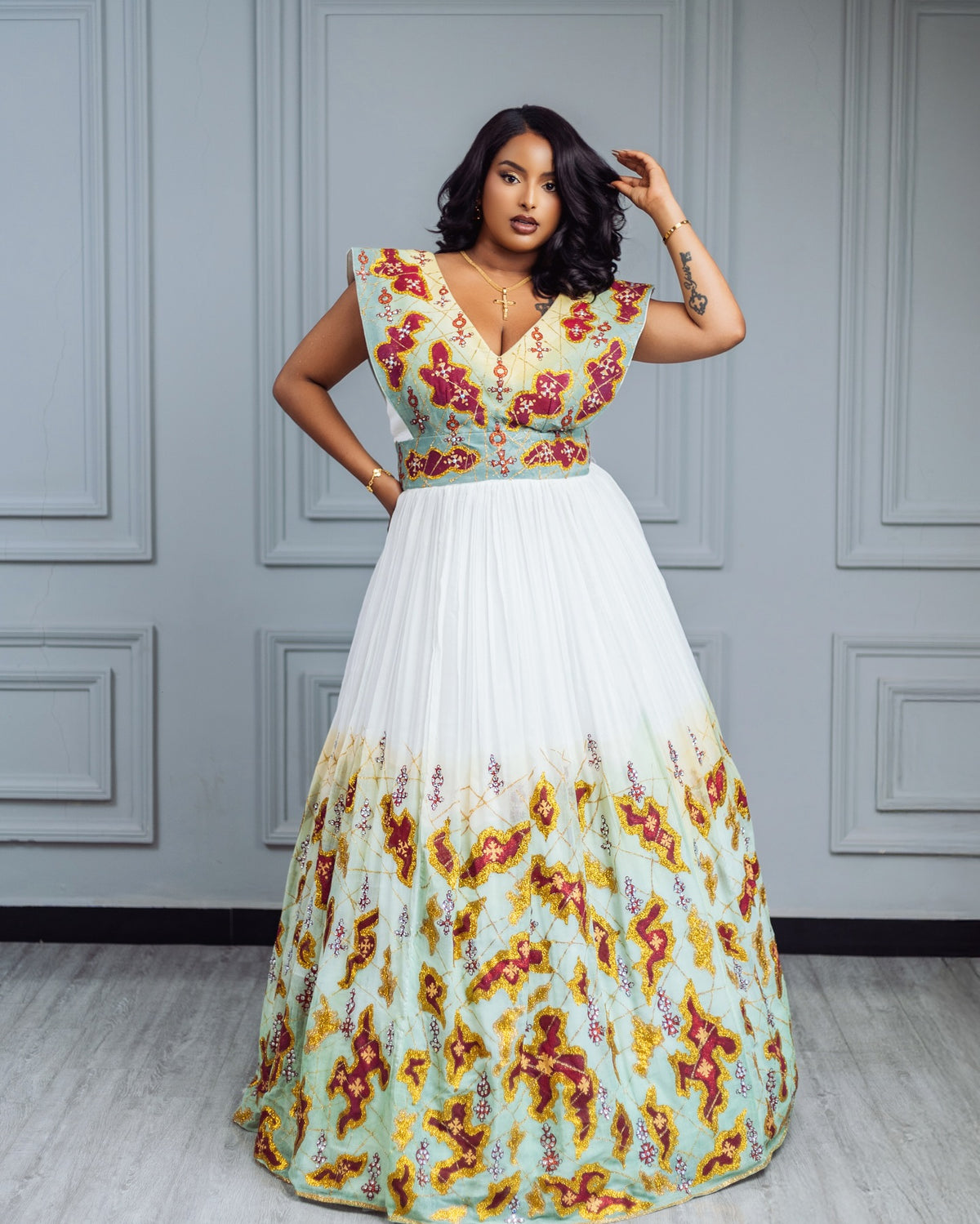 A woman modeling a floor-length formal habesha Wedding dress with a floral print. The dress features a light blue-green bodice and upper skirt with red and yellow floral patterns, transitioning into a solid white mid-skirt, set against a gray paneled wall.