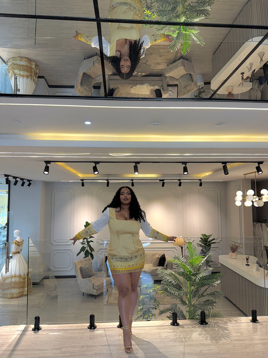 A woman posing in a modern, upscale showroom wearing a beige mini Habesha Wedding Dress  with yellow and white patterned trim and a white long-sleeve top. The background features a glass railing, indoor plants, and a mirrored ceiling showing an upside-down reflection of the scene.