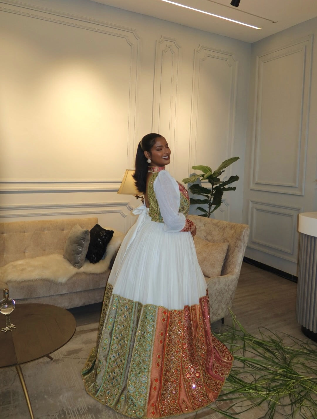 A woman wearing a traditional Indian lehenga Habesha Wedding Dress  with a white upper skirt, green embroidered bodice, and orange and green patterned bottom, standing in a decorated room with a sofa and plant.
