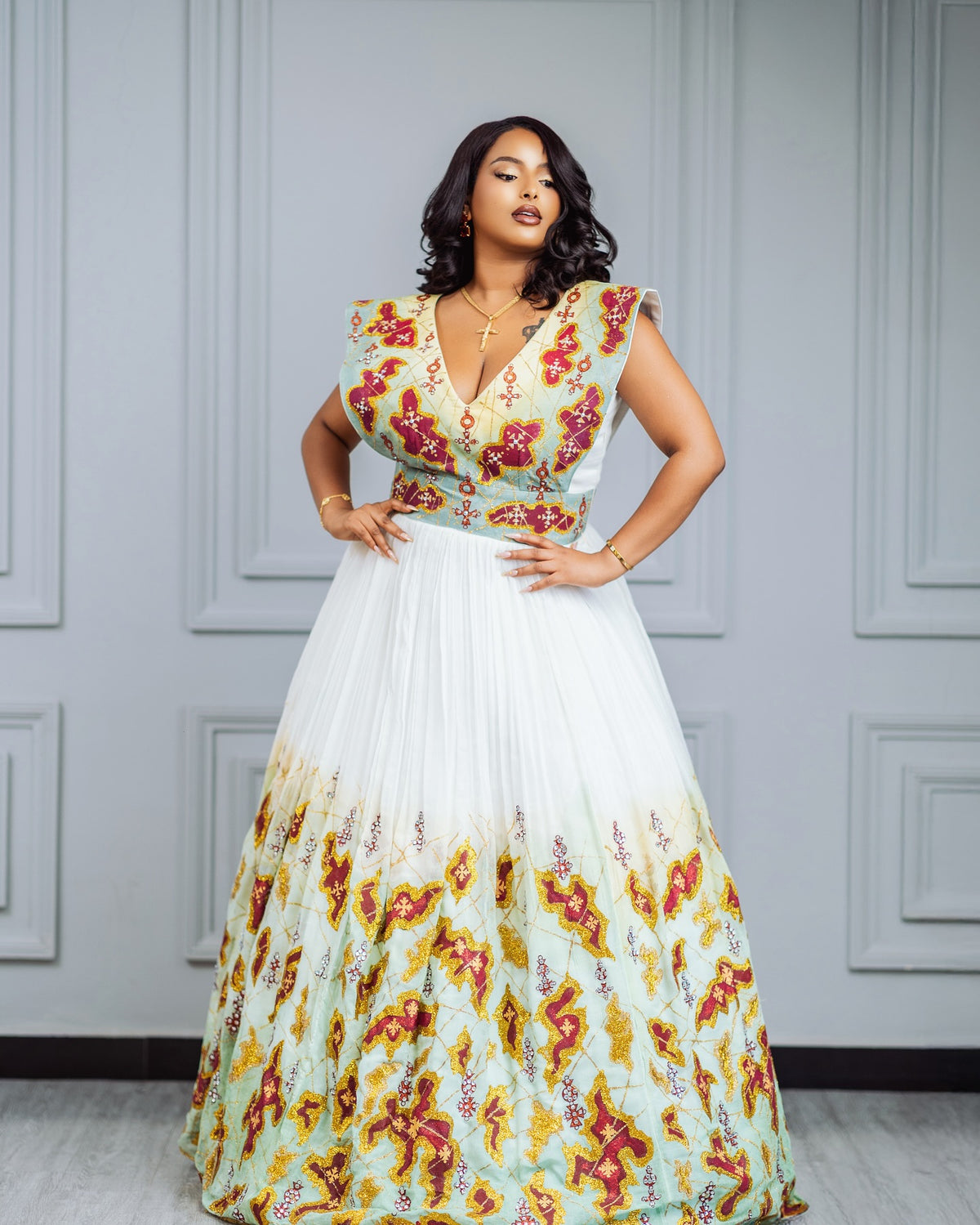 A woman models a sleeveless maxi habesha Wedding dress with a deep V-neckline, featuring a light green bodice with red and yellow floral embroidery and a flowing white skirt that fades into a matching floral print at the hem, set against a gray paneled wall.