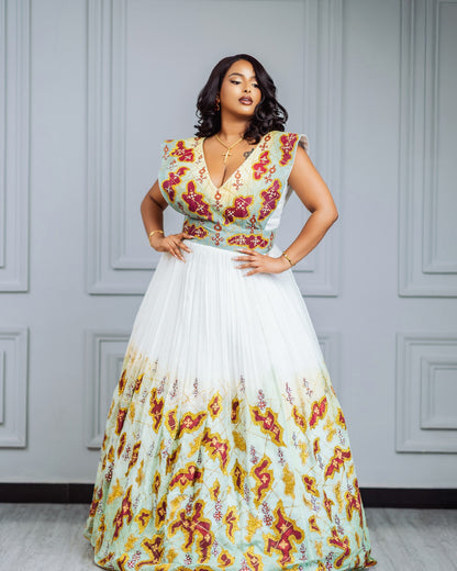 A woman models a sleeveless maxi habesha Wedding dress with a deep V-neckline, featuring a light green bodice with red and yellow floral embroidery and a flowing white skirt that fades into a matching floral print at the hem, set against a gray paneled wall.