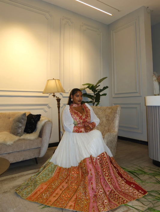 A woman wearing a traditional Habesha Wedding Dress  with a white pleated bodice and a highly embroidered, multicolored skirt in shades of orange, pink, and green, seated in a beige armchair inside a decorated room.