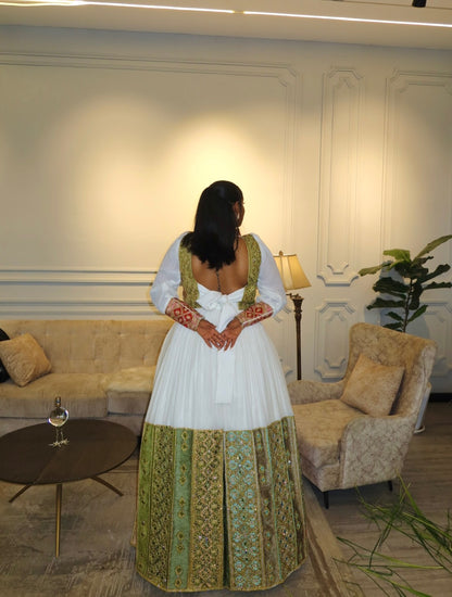 A woman stands in a living room wearing a traditional Habesha Wedding Dress  with ornate green and gold embroidered patterns on the skirt and sleeves. The dress features a low open back and a large bow at the center of the back.