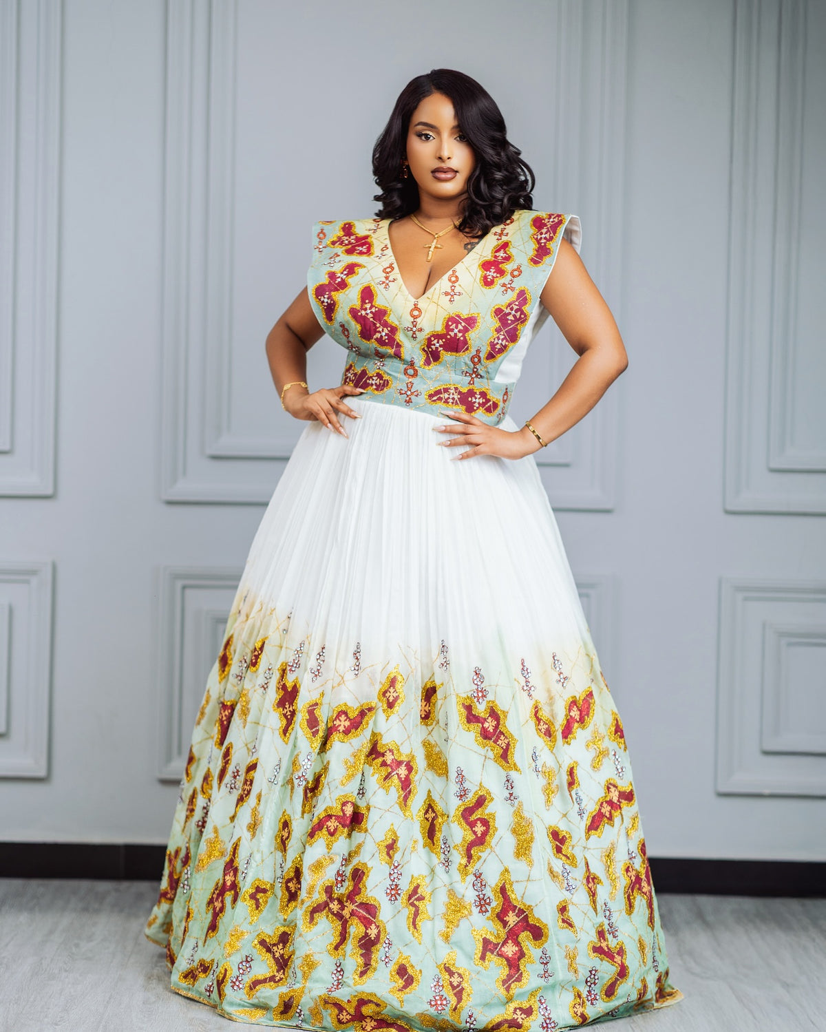 A woman models a sleeveless maxi habesha Wedding dress with a white tulle skirt and a floral printed bodice and lower skirt, standing against a gray paneled wall.