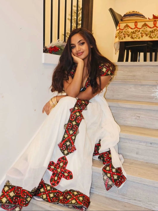 Woman in a white Ethiopian traditional dress  with red and gold embroidery sitting on stairs.