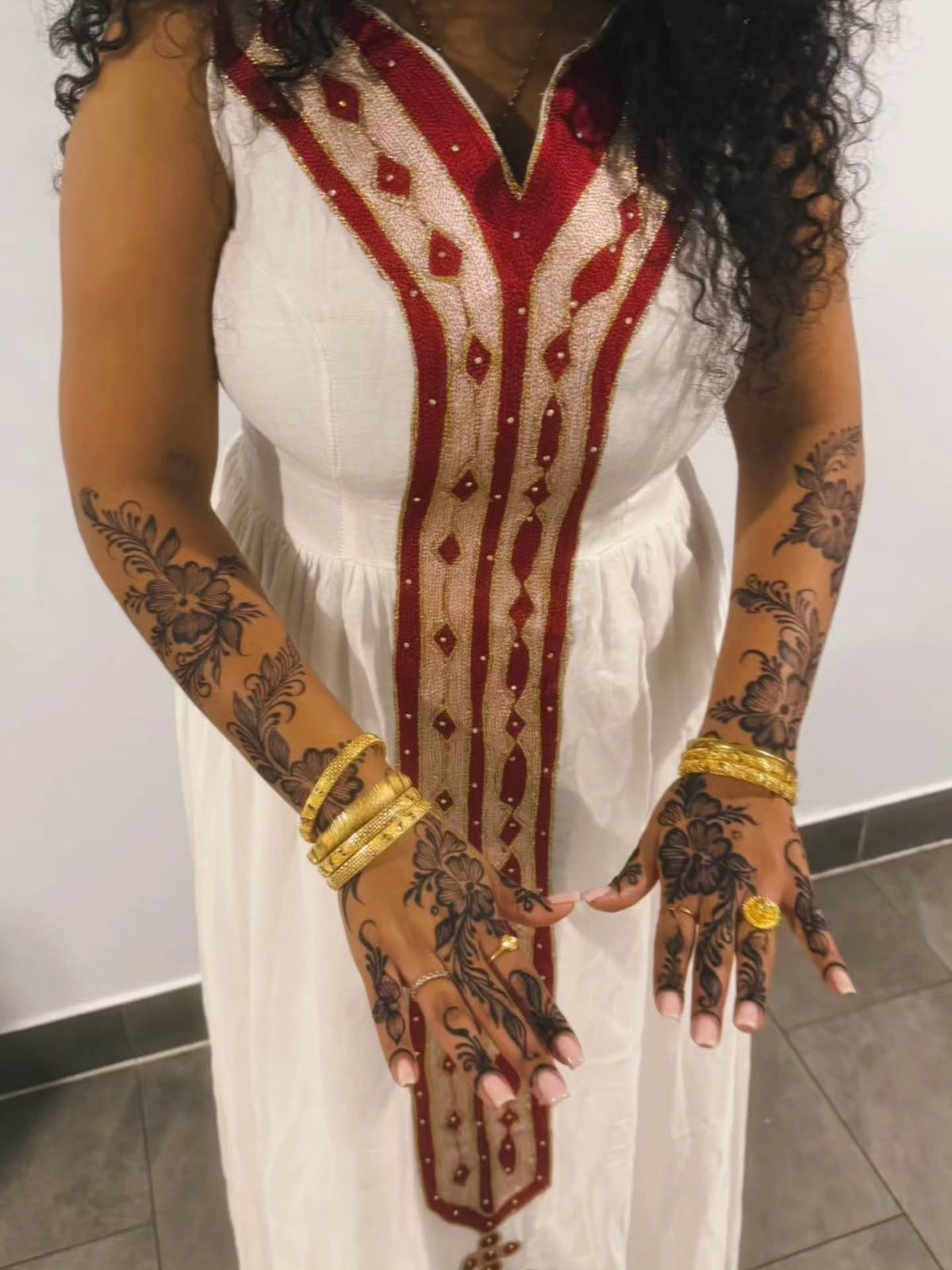 A woman wearing a white sleeveless Habesha Kemis  with a red and gold embroidered front placket, shown from the shoulders to the knees against a plain indoor wall.