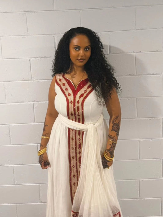 A woman with long curly black hair stands against a white painted brick wall wearing a sleeveless white Habesha Kemis  with red embroidered vertical panels and a matching white waist tie.