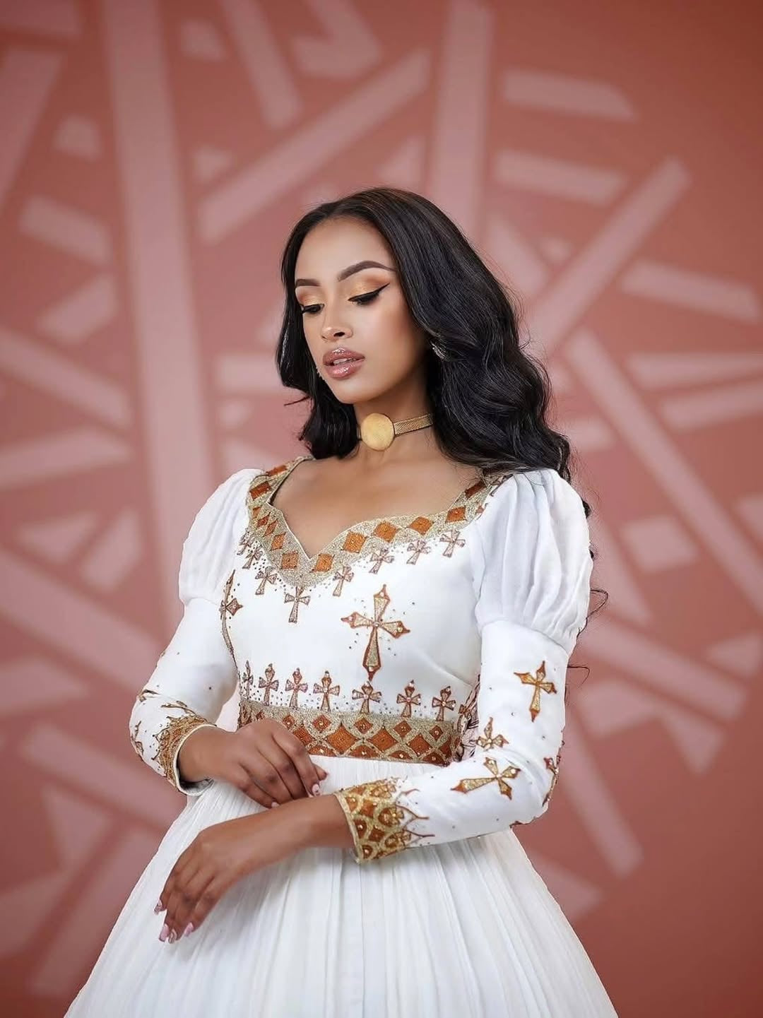 Woman wearing a white Habesha Dress with gold embroidery against a pink geometric patterned background