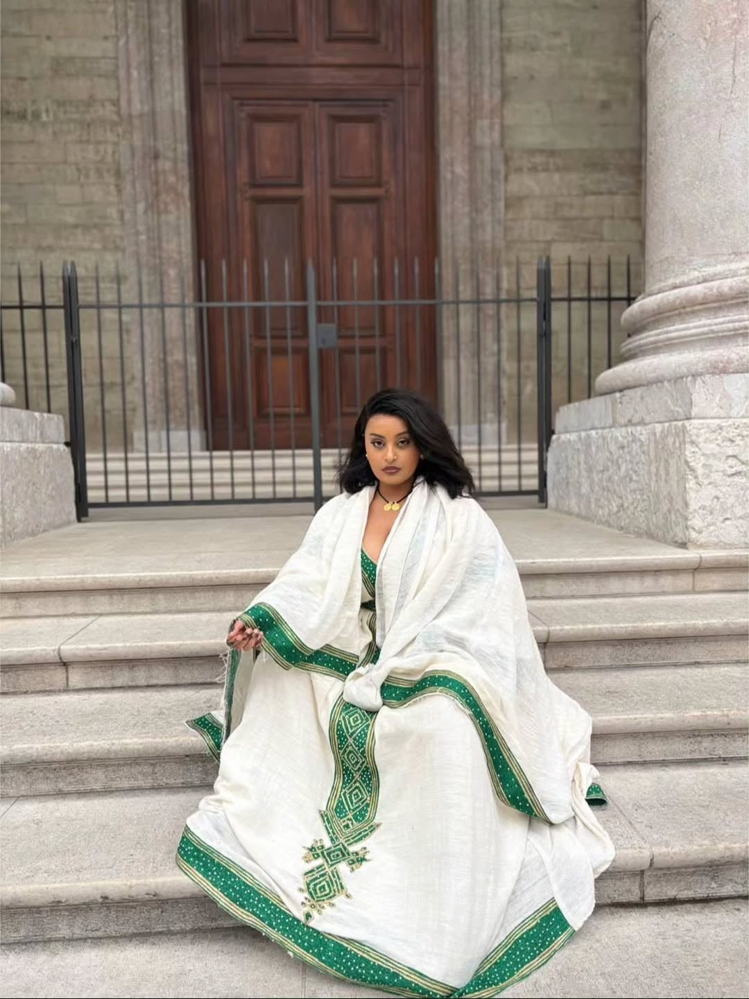 Woman in a white and green traditional Ethiopian Traditional Dress sitting on steps in front of a building.