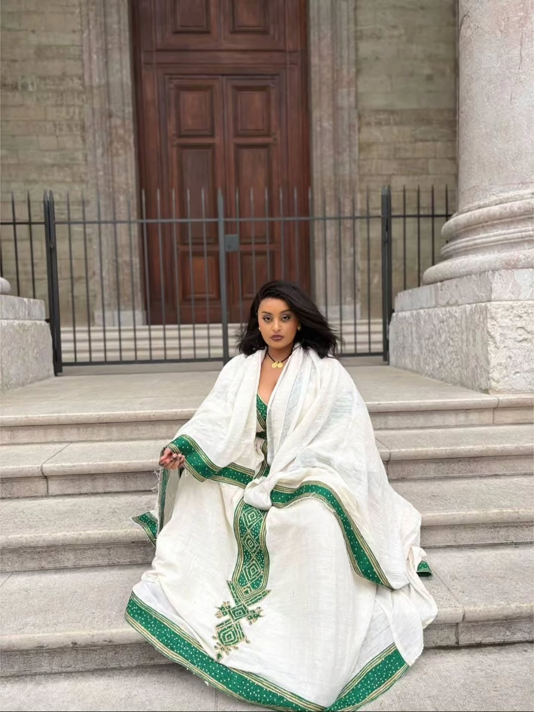 Woman in a white and green embroidered Ethiopian Traditional Dress sitting on steps in front of a building.