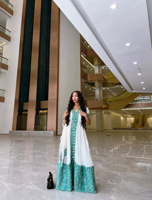 A woman modeling a traditional Habesha Kemis consisting of a white kurta with teal green geometric patterns, a matching dupatta, and wide-leg trousers. She is standing in a spacious, modern indoor lobby with polished marble floors and beige walls.