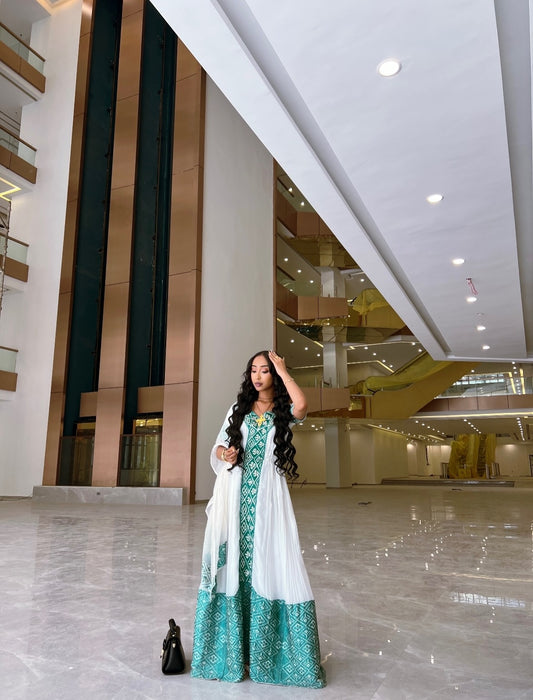 A woman models a traditional long Habesha Kemis  with a white base and teal geometric patterns, styled with a matching sheer white dupatta in a modern indoor setting.