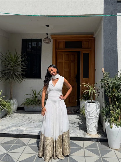 Woman in a white Habesha Dress  with gold trim standing in a courtyard with plants and a wooden door.