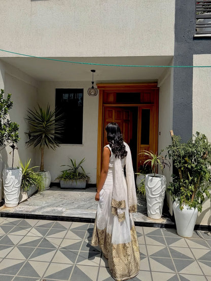 Woman in a white and gold traditional Habesha Dress  standing in front of a building entrance with plants around.
