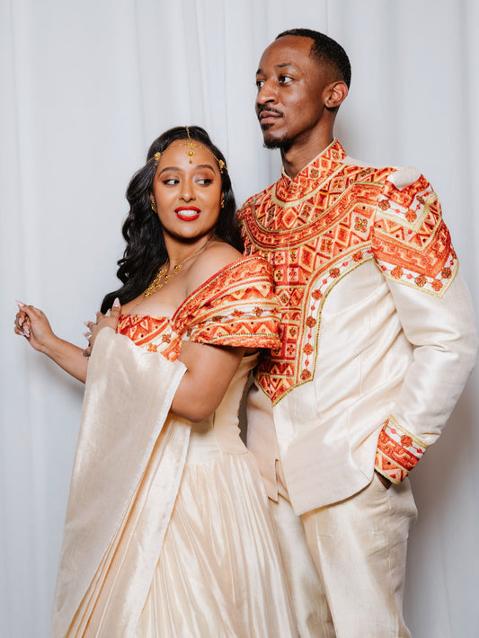 A couple posing in matching off-white traditional Ethiopian matching couple outfit with intricate orange and red embroidered patterns (tibeb).