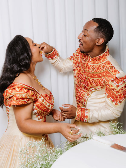 A couple posing in matching off-white traditional Ethiopian matching couple outfit with intricate orange and red embroidered patterns (tibeb).