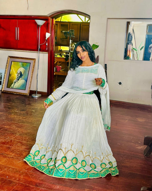A woman wearing a white traditional Habesha Kemis with green and gold embroidery, standing in an indoor room with wooden floors and home decor in the background.