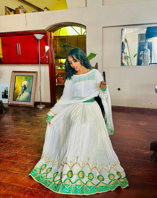 A woman models a white traditional Habesha Kemis with green and gold embroidery, featuring a flared skirt, a matching white dupatta, and a green waistband, set in an indoor room with wooden floors.