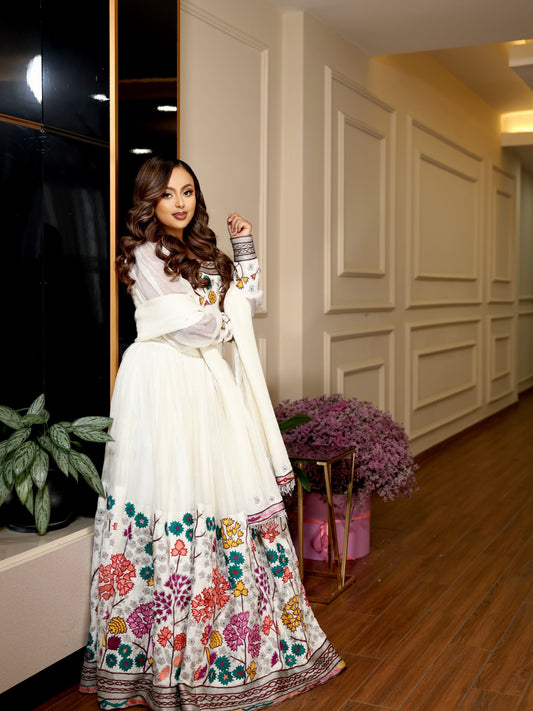 A woman models a traditional white embroidered Ethiopian Clothing with colorful floral patterns along the hem and sleeves, standing in an elegant indoor hallway with wooden floors and decorative plants.