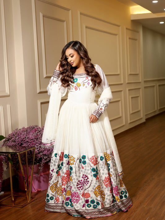 A woman models a traditional white Ethiopian Clothing with colorful floral embroidery concentrated along the hem and sleeves, set against an elegant indoor hallway with cream paneled walls and wooden floors.